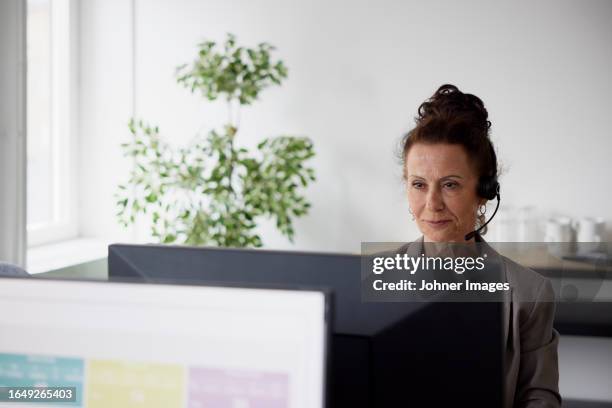 smiling mature businesswoman using headset in office in front of computer screen - office 60s computer person in front of screen stock pictures, royalty-free photos & images