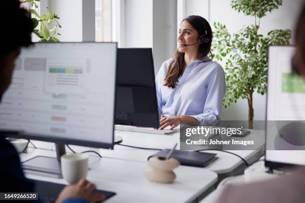 smiling mid adult businesswoman using headset in office in front of computer screen - office 60s computer person in front of screen stock pictures, royalty-free photos & images