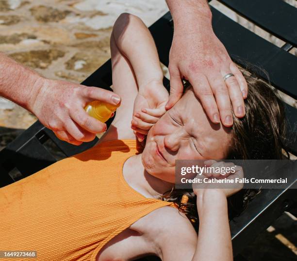 a little girl grimaces as a parent applies sunscreen to her face with a spray bottle - protection luxe stockfoto's en -beelden