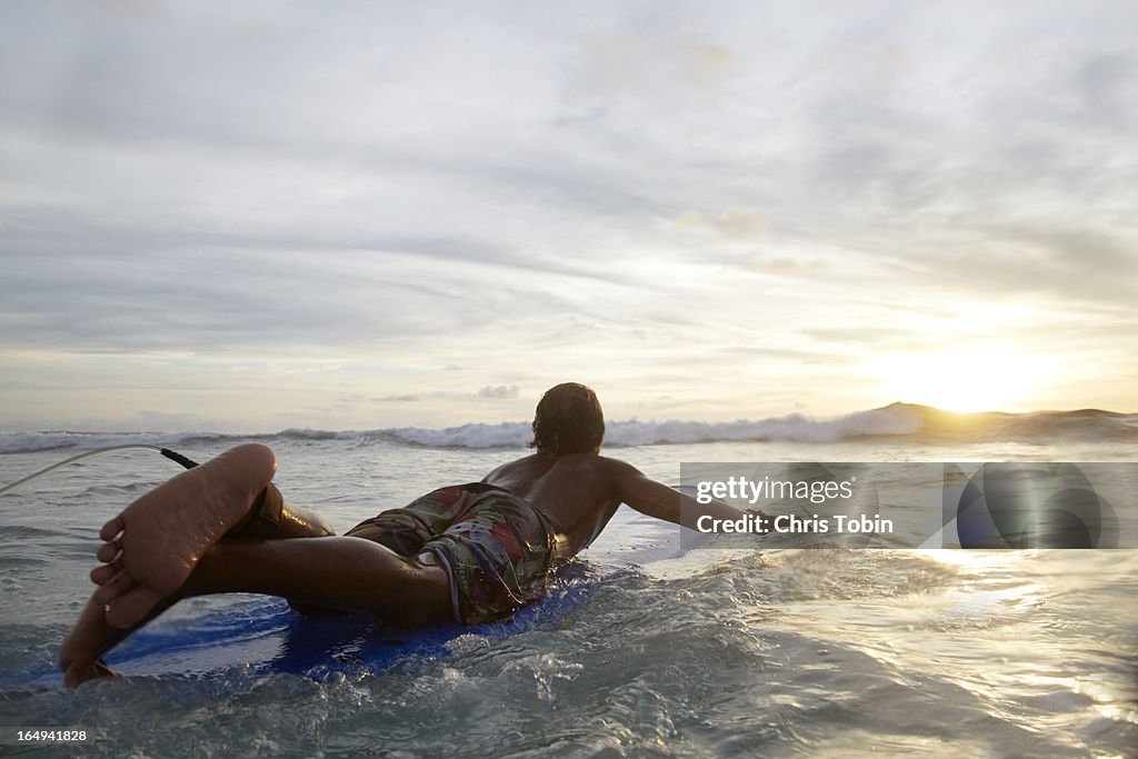 Guy paddling on surfboard into sunset