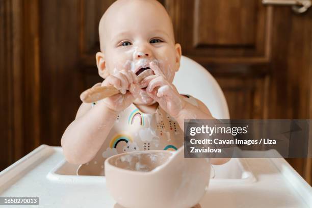 cute grubby six month old baby eating yogurt by himself - baby eating imagens e fotografias de stock