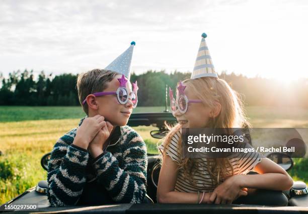 girl & boy sat in a convertible car with party hats and glasses - bell boy stock-fotos und bilder