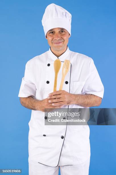 chef man in chef's suit and hat, holding a wooden spoon and fork, on blue background. concept for restaurant, cooking, menu, fast food, delicatessen and chef. - wooden spoon stock pictures, royalty-free photos & images