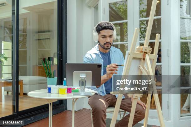 an asian-american mid-adult man spending quality time by taking an e-learning course on painting, and creating colorful watercolor pictures by watching on a digital tablet and using a headset - acrylic on canvas stock pictures, royalty-free photos & images