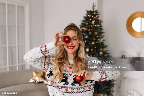 portrait of a young cheerful woman with red lips and curly hair in a knitted sweater laughing and holding a red christmas ball covering her eyes with it against the background of a christmas decorated christmas tree at a holiday in december at home - decorar fotografías e imágenes de stock