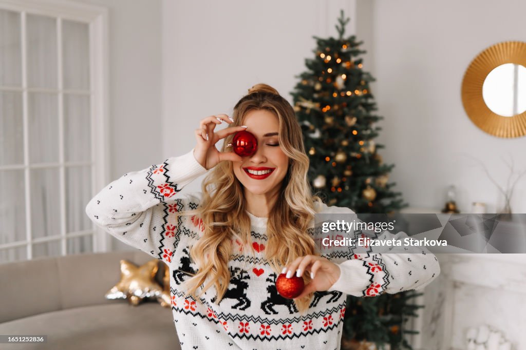 Portrait of a young cheerful woman with red lips and curly hair in a knitted sweater laughing and holding a red Christmas ball covering her eyes with it against the background of a Christmas decorated Christmas tree at a holiday in December at home