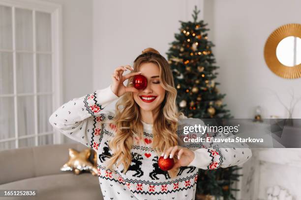 portrait of a young cheerful woman with red lips and curly hair in a knitted sweater laughing and holding a red christmas ball covering her eyes with it against the background of a christmas decorated christmas tree at a holiday in december at home - christmas fun stock pictures, royalty-free photos & images