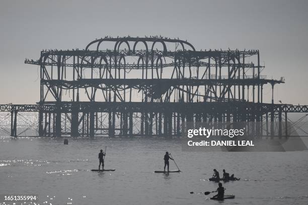 People paddleboard and canoe past the derelict west pier, enjoying the sun and the sea off the beach at Brighton, on the south coast of England on...