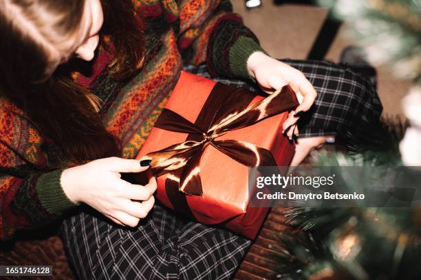 young woman tying ribbon while wrapping christmas present sitting on the couch at home - lint strik stockfoto's en -beelden