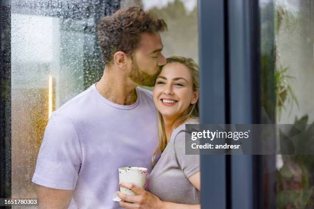 man kissing his girlfriend at home while looking through the window on a rainy day - couples kissing shower stockfoto's en -beelden