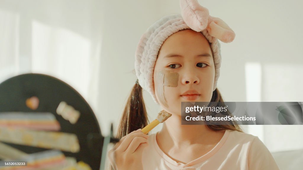 Happy mixed Asian preteen girl applying clay facial mask at home, skincare routine, self care and wellness concept