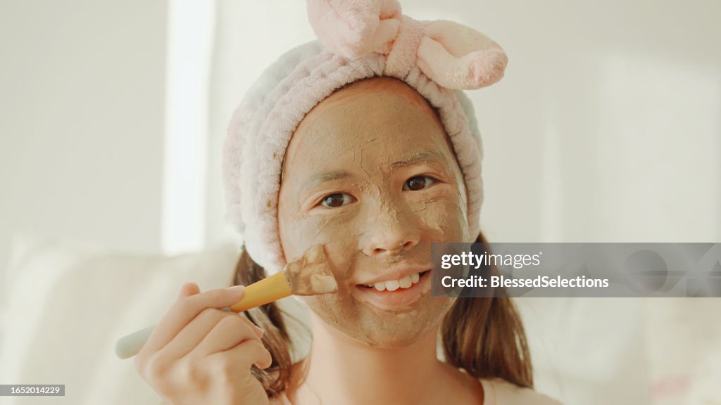 POV of Happy mixed Asian preteen girl applying clay facial mask at home