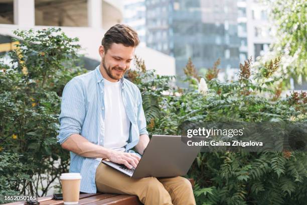 brunette young man in casual clothes working on laptop sitting on bench in the city - brunette sitting at desk stock-fotos und bilder