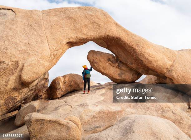 woman is standing under a large rock in joshua tree national park in southeastern california - joshua tree rock stock pictures, royalty-free photos & images