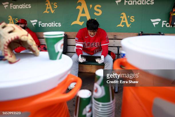 Shohei Ohtani of the Los Angeles Angels looks at a tablet while sitting in the dugout before their game against the Oakland Athletics at RingCentral...