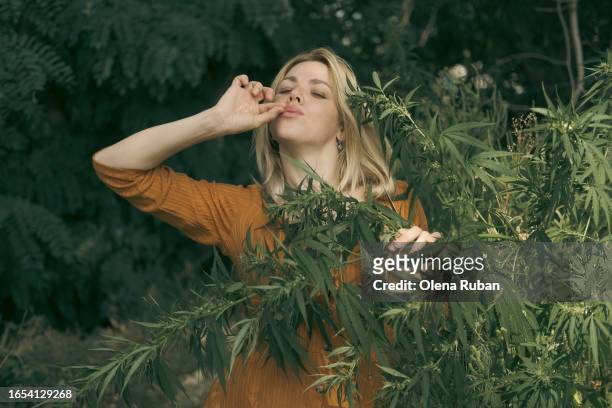 young woman holding fingers in smoking gesture near a hemp bush. - cannabinoid fotografías e imágenes de stock