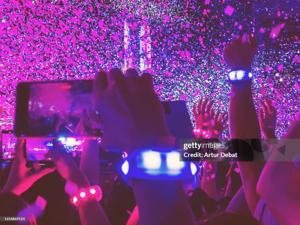 A group of people ecstatic with led bracelet recording with smartphone the concert with hands up, confetti and energy.