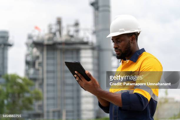 african america expert electrical engineer in safety suit and hardhat using a digital tablet to analyze plant data in the power plant factory. ensuring the plant's efficiency and safety is paramount. - premium gasoline stock pictures, royalty-free photos & images