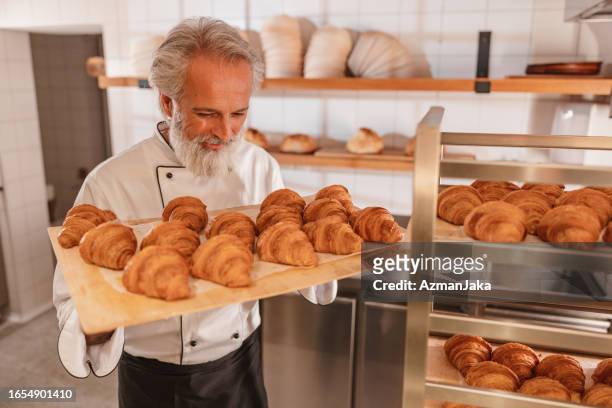 glücklicher reifer bäcker sucht und hält ein tablett voller frisch gebackener croissants - baker smelling bread stock-fotos und bilder
