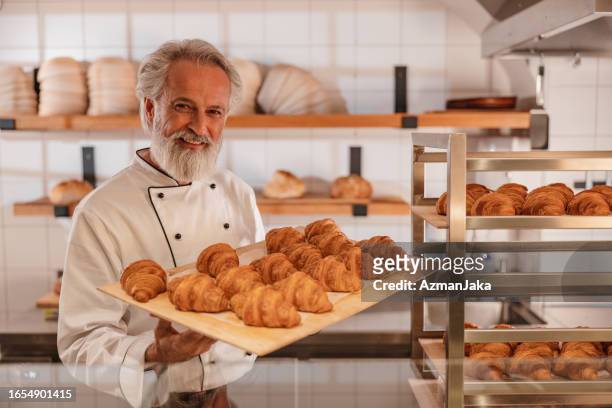 smiley kaukasischer bäcker hält ein tablett voller frisch gebackener croissants - baker smelling bread stock-fotos und bilder