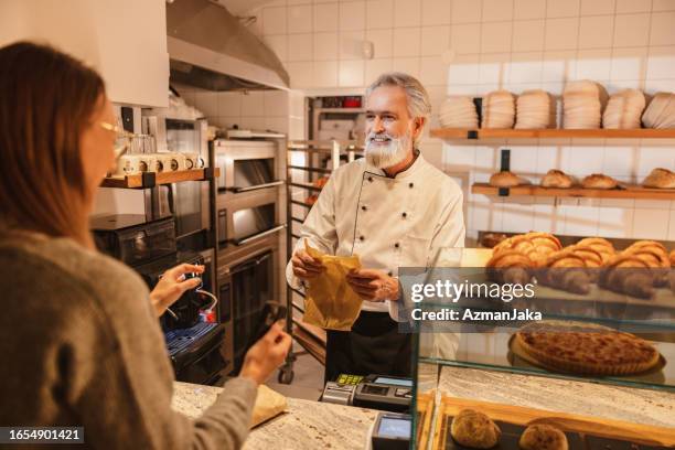 mature baker giving an order to a young costumer at the bakery - baker smelling bread stockfoto's en -beelden