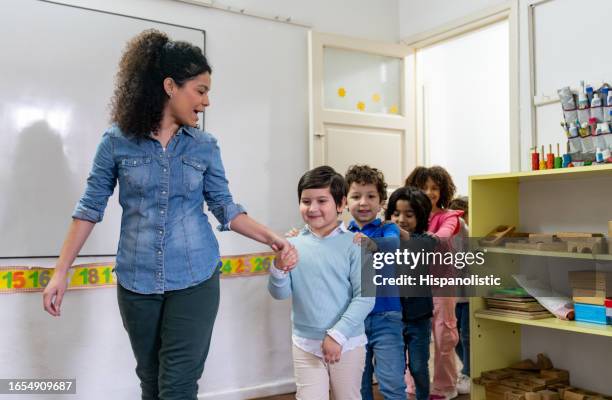 cheerful teacher walking into the classroom with her students all following in a row with hands on their classmates shoulders and smiling - kids in a row stock pictures, royalty-free photos & images