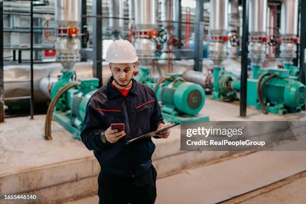 ein junger mann wird gezeigt, wie er mit seinem mobiltelefon mit einem kollegen im heizwerk spricht - boiler engineer stock-fotos und bilder