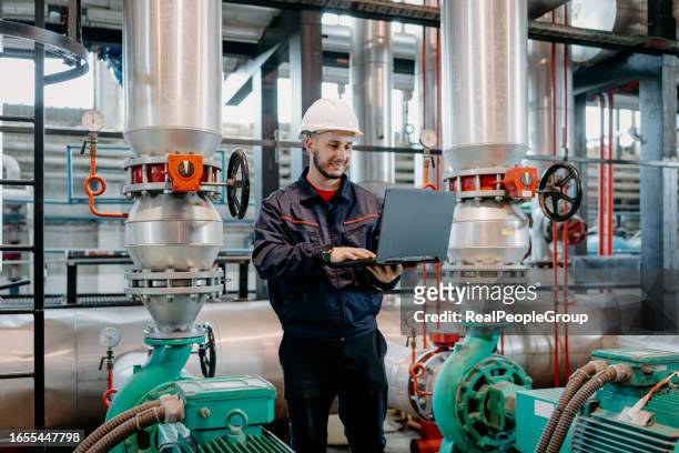 junger mann, der seinen laptop benutzt, um die systeme des heizwerks effizient zu verwalten - district heating plant stock-fotos und bilder