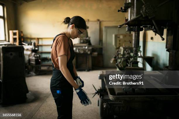 woman worker putting on safety gloves standing at drill machine in metal workshop - leather glove stock pictures, royalty-free photos & images