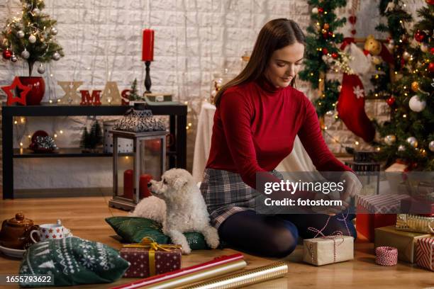 young woman sitting on the floor and tying a string around a christmas gift box she is decorating - candy wrapper stock pictures, royalty-free photos & images