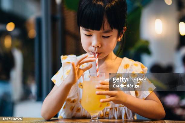 cute little asian girl enjoying a glass of iced apple juice with a paper straw in cafe. sustainable lifestyle. environmental conservation concept - vruchtensap stockfoto's en -beelden