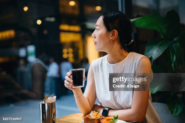 young asian woman sitting inside a cozy cafe, having a cup of coffee. she is looking at view through the window. enjoying a relaxing moment on a fresh morning. people, food and lifestyle - me time stock pictures, royalty-free photos & images