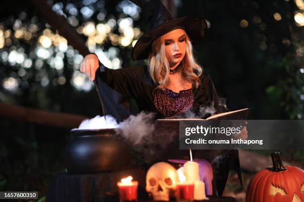 the fantasy woman witch conjures, holds a magic wand and a magic book in her hands, and reads spell white magic smoke from boiling vat. halloween theme. witch performs ceremony on halloween day - magic wand stockfoto's en -beelden
