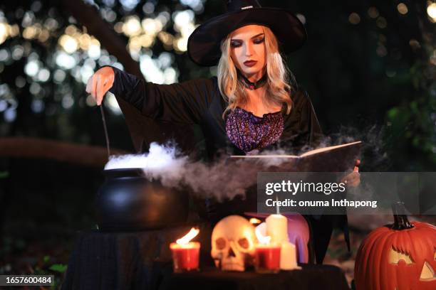 the fantasy woman witch conjures, holds a magic wand and a magic book in her hands, and reads spell white magic smoke from boiling vat. halloween theme. witch performs ceremony on halloween day - witch wand stock pictures, royalty-free photos & images