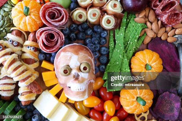 full frame image of cheese-charcuterie board, prosciutto di parma skull, salami roses, cucumber, cheddar cubes, cherry tomatoes, almonds, apricots, blueberries, sausage mummies, satsuma pumpkins, pretzels, beetroot crisps, elevated view - frankenstein stock pictures, royalty-free photos & images