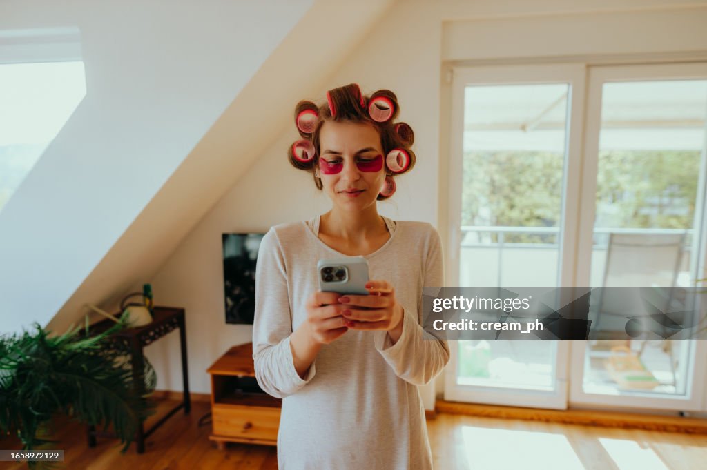 Woman with hair rollers and eye mask using a smartphone at home