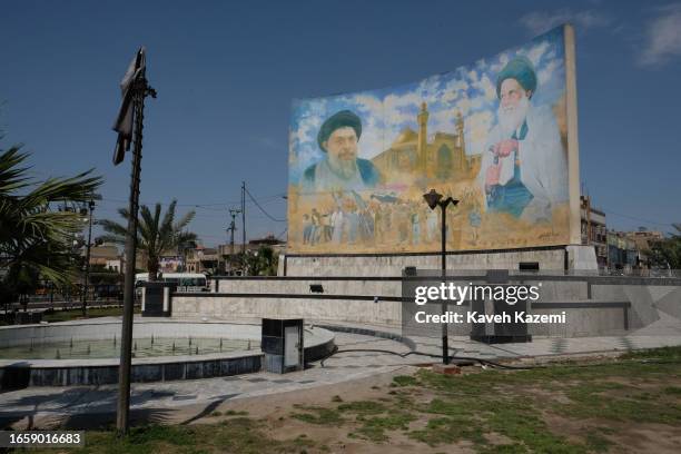 Children sit by a pond with their feet in water, in front of a huge billboard showing pictures of deceased Grand Iraqi Ayatollahs Muhammad al-Sadr...