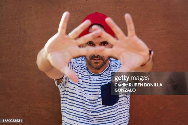 young latino man wearing red cap and looking at the camera while gesturing to stop with two hands on brown background. open palm. self defending concept. - gonzalo caballero fotografías e imágenes de stock