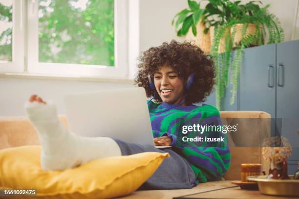 young woman with broken leg using laptop at home - african cast broken leg stockfoto's en -beelden