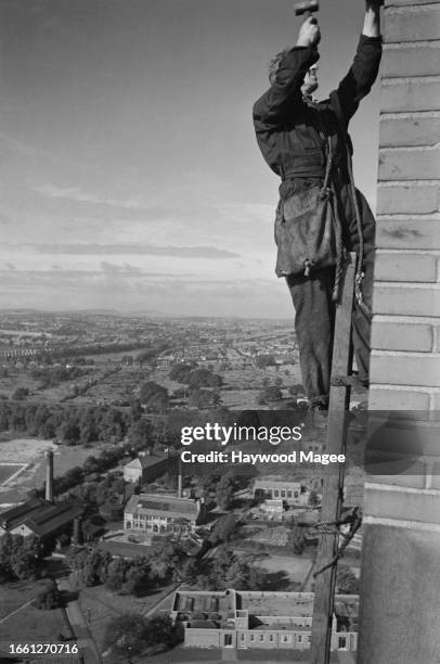Steeplejack at work with a hammer on Birmingham University's clock tower, high above the countryside and factory buildings, 1942. Original...
