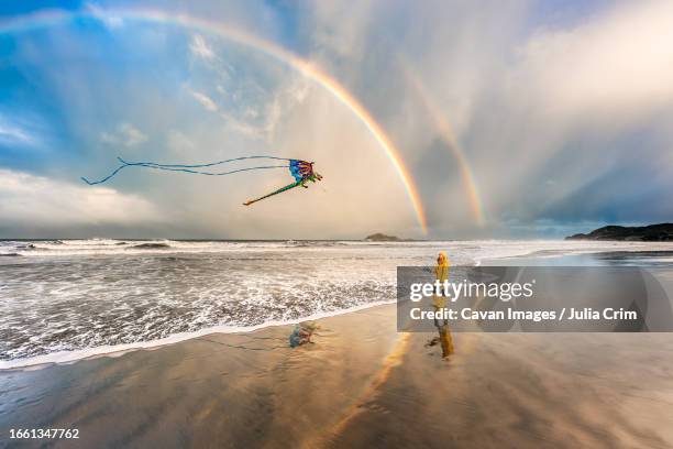 girl in yellow coat flying kite on beach with double rainbow - double rainbow stock pictures, royalty-free photos & images