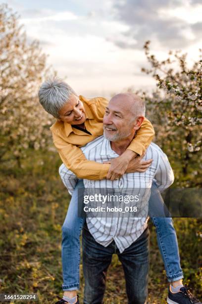 active senior man carrying his wife piggyback on sunny day in nature - spring romance stock pictures, royalty-free photos & images