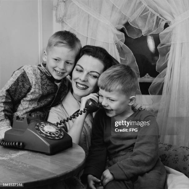 Woman sits with two young boys, all smiling as she holds a telephone receiver to the ear of one of the boys, United States, circa 1955.