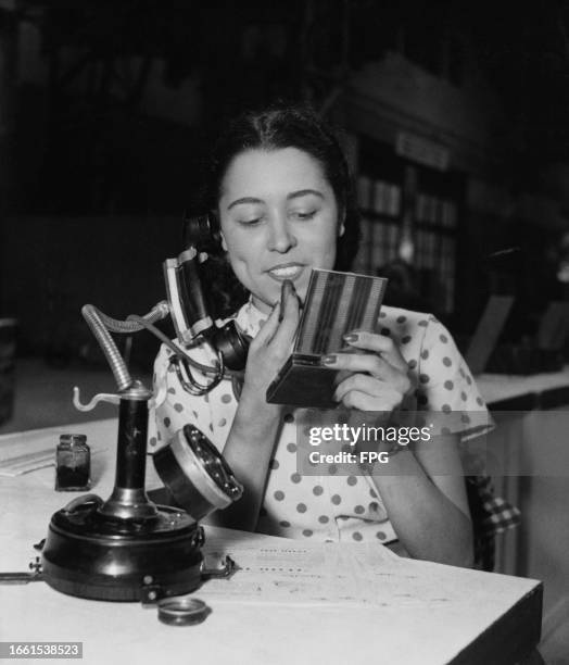 Woman applies lipstick with her right hand, holding a mirror in her left, while speaking on the phone with the receiver on an adjustable arm, during...