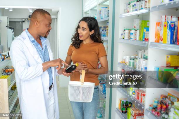 professional african american pharmacist man with skinhead and beard helping beautiful latin woman patient who has stomachache with medicine recommendation at drug store. mid adult latin pharmacist advising, talking, and explaining indication of drug. - latin american and hispanic shopping bags stockfoto's en -beelden