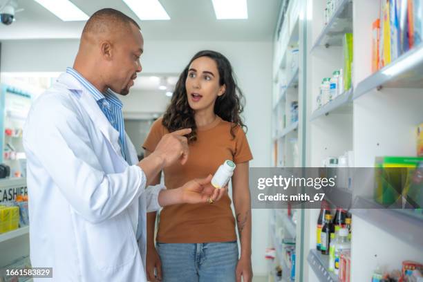 latin american woman buying medicines at the drugstore with the assistance of the professional pharmacist male. mid adult african american woman shopping in the pharmacy and listening to pharmacist for the suggestion and recommendation. - latin american and hispanic shopping bags stockfoto's en -beelden