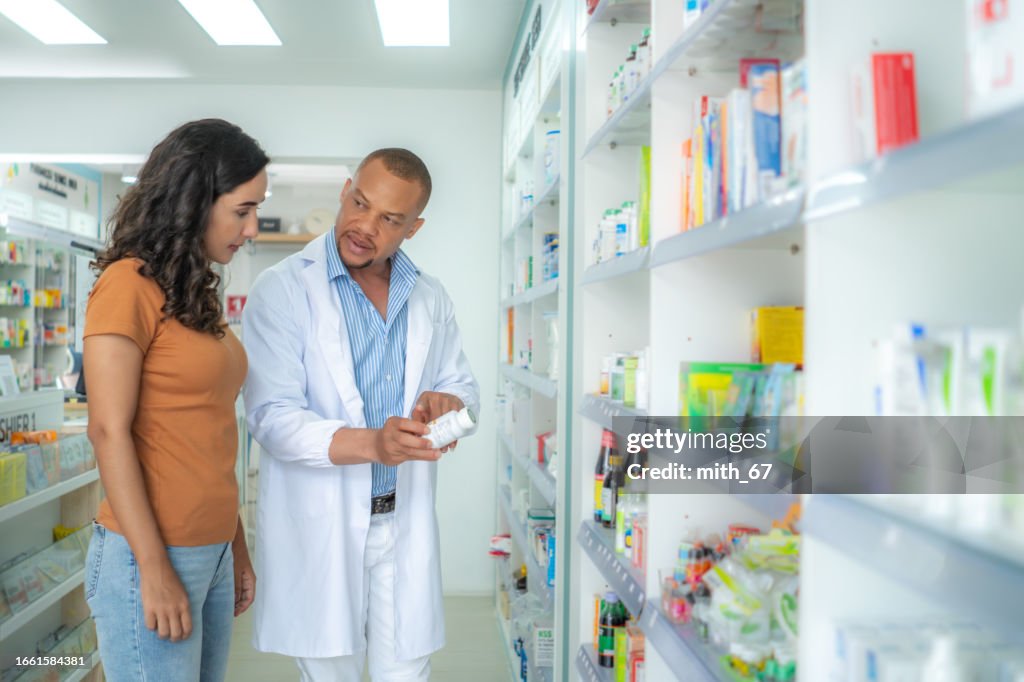 Latin mid adult female customer listening, talking to professional black pharmacist male at drug store. Mid adult African American pharmacist male with skinhead and beard
explaining medication following the instruction from label to customer.