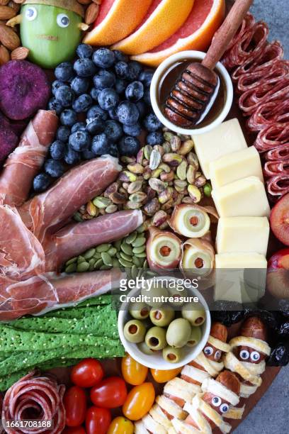 full frame image of cheese-charcuterie board, prosciutto di parma hand, pink grapefruit, salami roses, cheddar cubes, cherry tomatoes, frankenstein monster kiwi fruit, almonds, sausage mummies, blueberries, satsuma pumpkins, grey background, elevated view - frankenstein stock pictures, royalty-free photos & images
