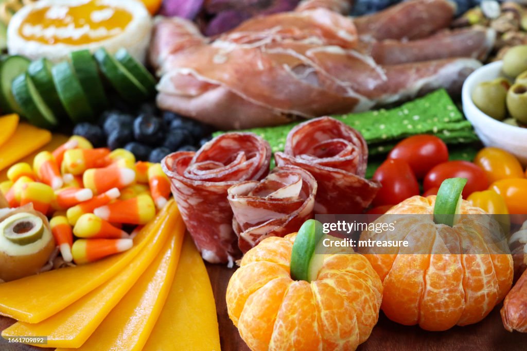 Immagine a pieno formato di tagliere di formaggi-salumi di Halloween, brie di zucca, mano di prosciutto di Parma, zucche satsuma, cetrioli, formaggio rosso Leicester, rose di salame, mirtilli, cracker, mais caramellato, pistacchi, fette di mela verde, mess