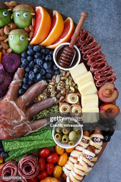 image of cheese-charcuterie board, prosciutto di parma hand, pink grapefruit, salami roses, cheddar cubes, cherry tomatoes, frankenstein monster kiwi fruit, almonds, sausage mummies, blueberries, satsuma pumpkins, grey background, elevated view - frankenstein stock pictures, royalty-free photos & images